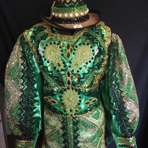 May include: A green and gold costume with intricate beading and embroidery. The costume features a long-sleeved shirt with a high collar and a matching skirt. The shirt has a heart-shaped design on the front, and the skirt has a geometric pattern. The costume is accented with black trim and a straw hat with a black band.