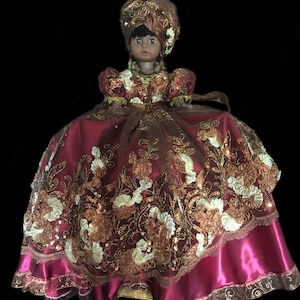 May include: A decorative doll with dark skin, wearing a burgundy satin dress with gold and white floral embroidery. The doll has a matching turban and is adorned with gold accents and sequins. The doll is set against a black background.