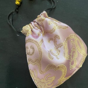 Op de afbeelding: Een klein roze trekkoordtasje met een gouden bloemenpatroon en gouden trekkoordsluitingen.