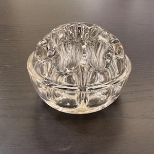 May include: A clear glass flower frog with a rounded base and a domed top. The top has multiple holes for arranging flower stems. The glass has a textured, ribbed design. The frog is sitting on a dark surface.
