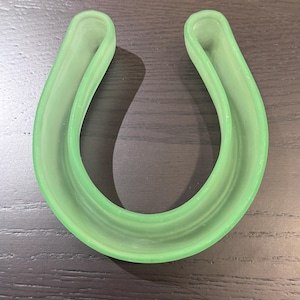 May include: A light green, U-shaped dental appliance, likely a mouth guard or retainer. The object is made of a translucent material and has a smooth, curved design. The item is resting on a dark wooden surface.