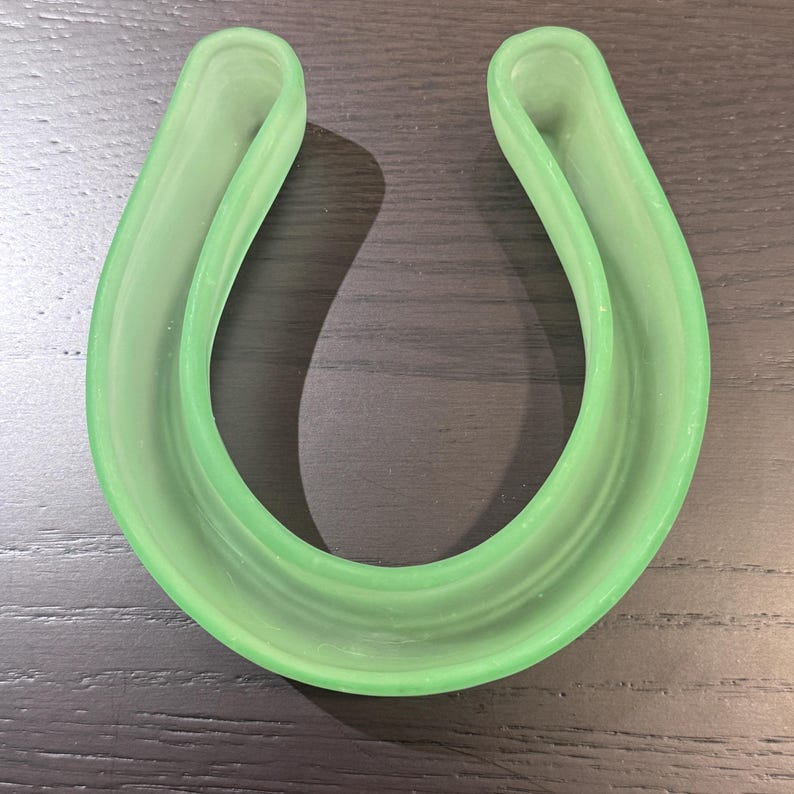 May include: A light green, U-shaped dental appliance, likely a mouth guard or retainer. The object is made of a translucent material and has a smooth, curved design. The item is resting on a dark wooden surface.