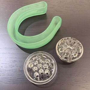 May include: An assortment of vintage glass flower arranging tools. Includes a light green curved frog, and two clear glass flower frogs with multiple holes for arranging stems. The tools are on a dark wood surface.