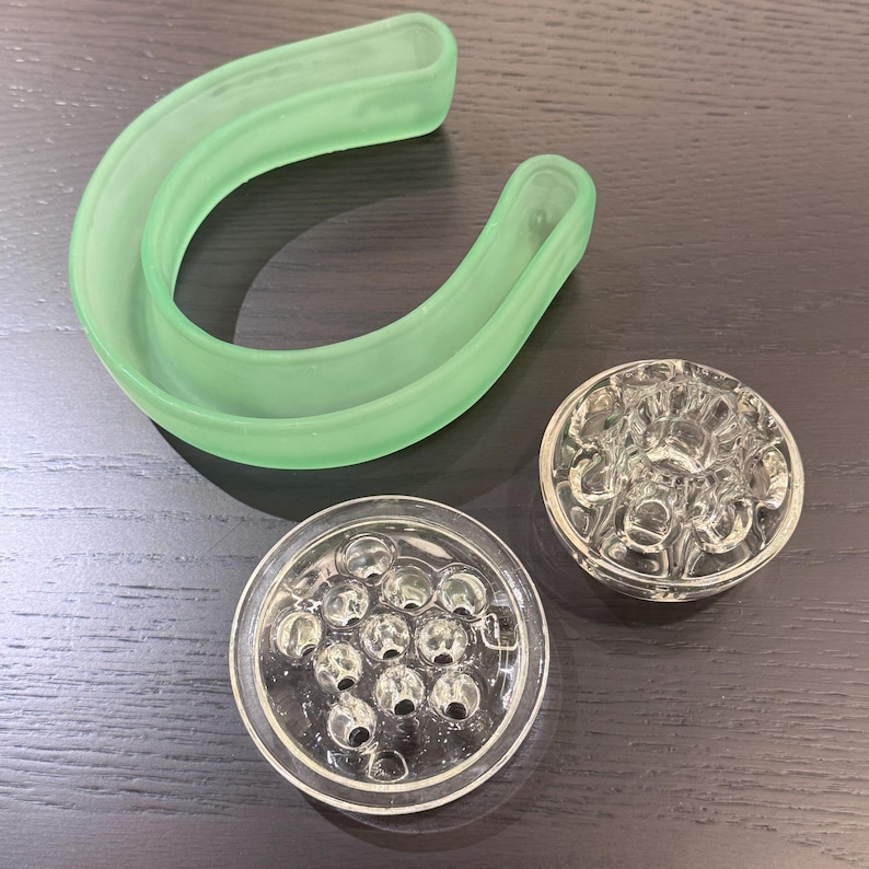 May include: An assortment of vintage glass flower arranging tools. Includes a light green curved frog, and two clear glass flower frogs with multiple holes for arranging stems. The tools are on a dark wood surface.
