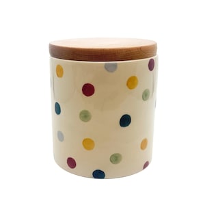 May include: A cylindrical ceramic canister with an off-white base featuring a scattered pattern of multi-colored polka dots in shades of red, yellow, blue, green, and grey. The container is topped with a flat, circular, natural wood lid.