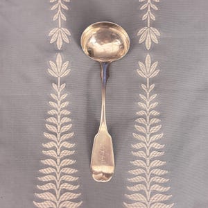Pode incluir: Uma concha de prata com uma tigela redonda e um cabo longo. A alça tem um design decorativo e uma pequena inscrição. A concha é colocada sobre um fundo azul claro com um padrão floral branco repetido.