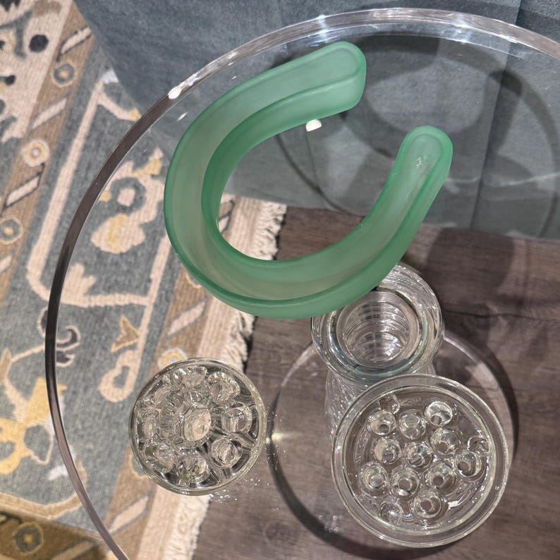 May include: A clear glass table with a light green, curved glass object and several clear glass flower frogs. The flower frogs have various hole patterns. The table is on a patterned rug.