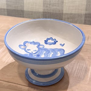 May include: A white ceramic bowl with a blue rim and base, featuring a hand-painted sheep design. The bowl has a pedestal base and is decorated with blue stripes and a sheep illustration. The bowl is suitable for serving food.