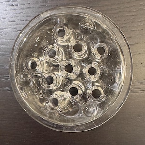 May include: Clear glass flower frog with multiple circular holes. The frog is round with a shallow dish shape and is designed to hold flower stems in place. The holes are of varying sizes and are evenly spaced.