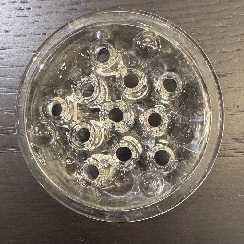 May include: Clear glass flower frog with multiple circular holes. The frog is round with a shallow dish shape and is designed to hold flower stems in place. The holes are of varying sizes and are evenly spaced.