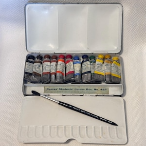 May include: An open, white metal Reeves' Students' Colour Box with a selection of watercolor paint tubes in various colors. A paintbrush with a black handle and squirrel hair bristles rests on the white palette. The box is labeled "Reeves' Students' Colour Box No. 42P."