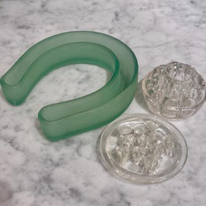 May include: Three floral arranging tools: a light green, curved, trough-like piece, and two clear glass flower frogs. One frog is a shallow, round disc with holes, and the other is a small, round, bowl-shaped frog.