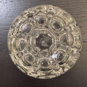 May include: A clear, circular glass knob with a textured, bubble-like design. The knob features a central indentation surrounded by larger, circular bubbles and smaller, irregular bubbles. The glass is transparent, allowing light to pass through, and is likely used for furniture or decorative purposes.