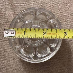 May include: Clear glass knob with a circular design and oval indentations. A yellow measuring tape shows the knob is approximately 2.5 inches in diameter. The knob is likely for a cabinet or drawer.