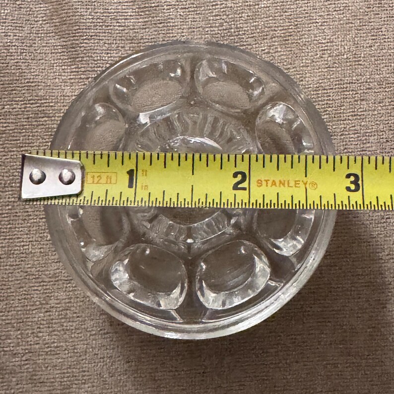 May include: Clear glass knob with a circular design and oval indentations. A yellow measuring tape shows the knob is approximately 2.5 inches in diameter. The knob is likely for a cabinet or drawer.