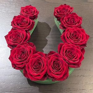 May include: A horseshoe-shaped floral arrangement featuring vibrant red roses nestled in a light green container. The roses are fully bloomed, showcasing their rich color and intricate petal arrangement. The arrangement sits on a dark wooden surface.