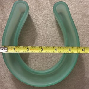 May include: A translucent, light green horseshoe-shaped object. The object is approximately 6 inches wide. A yellow measuring tape is placed across the opening of the horseshoe, showing the width.