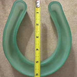 May include: A translucent, light green horseshoe-shaped object. A yellow tape measure is placed next to the object, showing a height of approximately 5.5 inches. The object has a smooth, rounded shape.