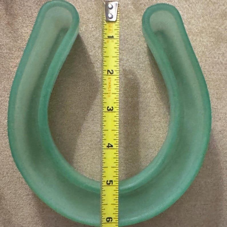 May include: A translucent, light green horseshoe-shaped object. A yellow tape measure is placed next to the object, showing a height of approximately 5.5 inches. The object has a smooth, rounded shape.