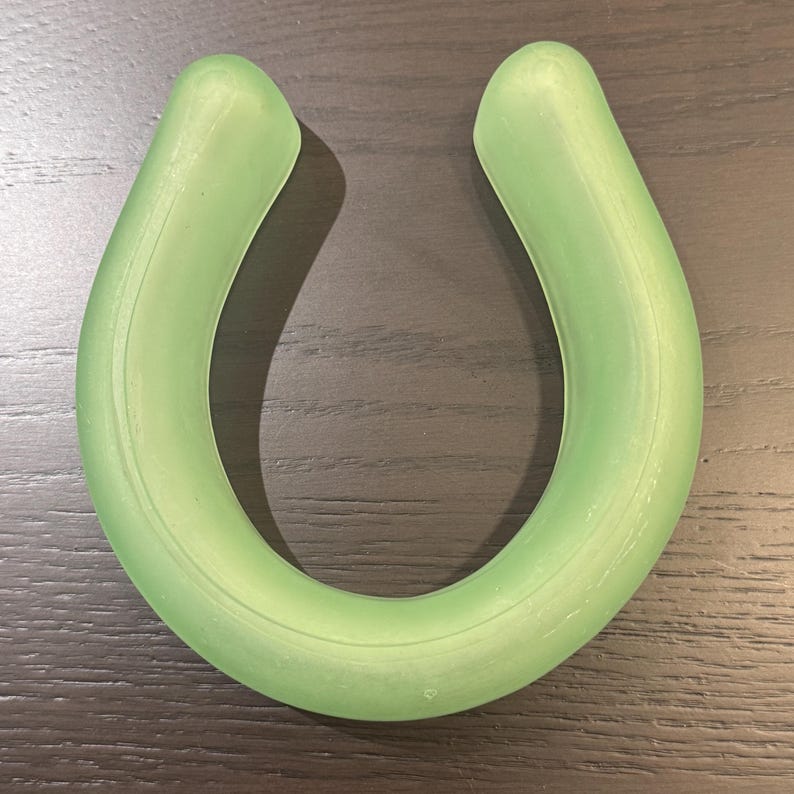 May include: A light green, U-shaped object with a smooth, rounded design. The object is made of a translucent material and appears to be a single piece. The item is resting on a dark wood surface.