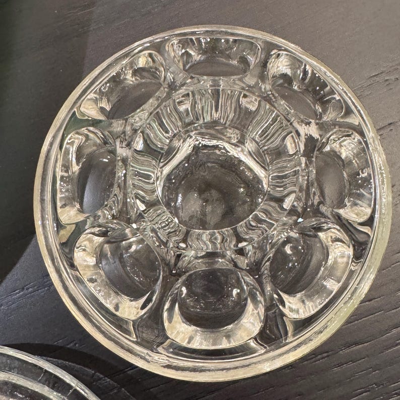 May include: A clear, circular glass coaster with a unique design. The coaster features a central depression surrounded by several circular indentations. The glass has a textured appearance, and the overall design is symmetrical.