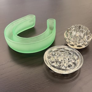 May include: An assortment of vintage glass flower frogs and a green glass crescent-shaped vase. The flower frogs are clear, with one round and one oval design. The green vase is a light, frosted green color.