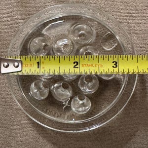 May include: Clear glass flower frog with multiple holes, approximately 3 inches in diameter. The flower frog is round with a decorative edge and is designed to hold flower stems in place.