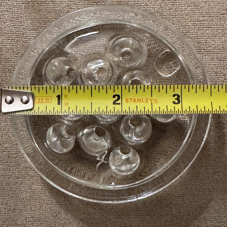 May include: Clear glass flower frog with multiple holes, approximately 3 inches in diameter. The flower frog is round with a decorative edge and is designed to hold flower stems in place.