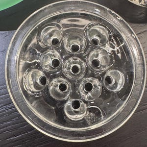 May include: Clear glass flower frog with a circular design. The frog has a series of holes arranged in a pattern, designed to hold flower stems in place. The glass is transparent, allowing a clear view of the stems.