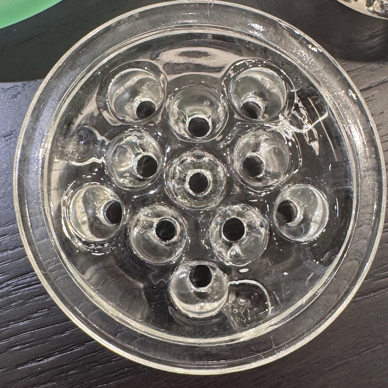 May include: Clear glass flower frog with a circular design. The frog has a series of holes arranged in a pattern, designed to hold flower stems in place. The glass is transparent, allowing a clear view of the stems.