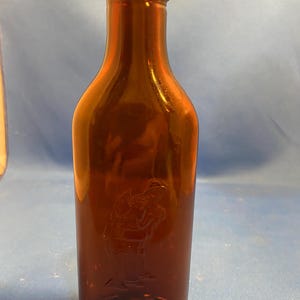 May include: An amber-colored glass bottle with a narrow neck and a threaded top. The body of the bottle features a subtle, embossed graphic of a figure standing in profile, integrated into the smooth, translucent surface of the glass material.