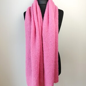 May include: A bright pink, knitted scarf draped over a black mannequin. The scarf is made of a soft, textured material and is displayed against a plain white background. The scarf is a solid color and appears to be hand-knitted.