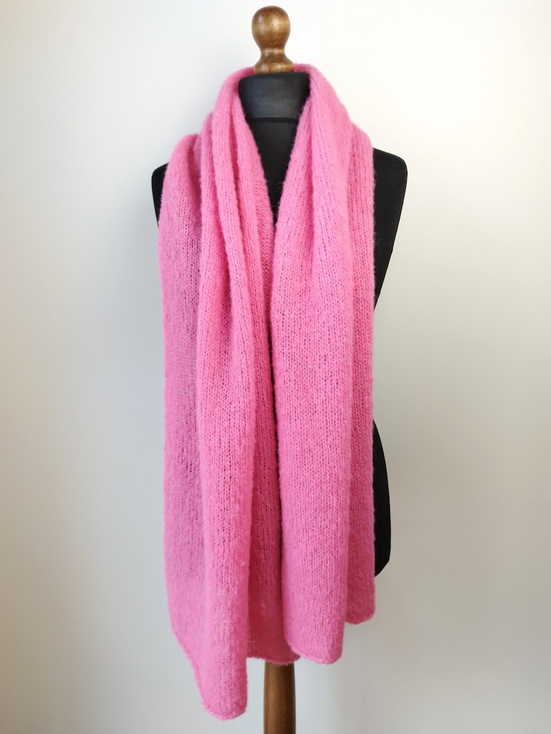 May include: A bright pink, knitted scarf draped over a black mannequin. The scarf is made of a soft, textured material and is displayed against a plain white background. The scarf is a solid color and appears to be hand-knitted.