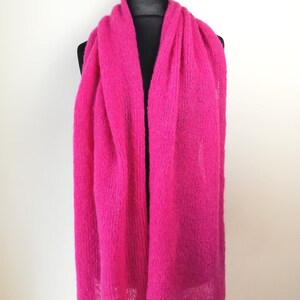 May include: A vibrant, hot pink scarf displayed on a black mannequin head and wooden stand. The scarf is made of a soft, knitted material and drapes down in long, flowing folds. The background is a plain white wall.
