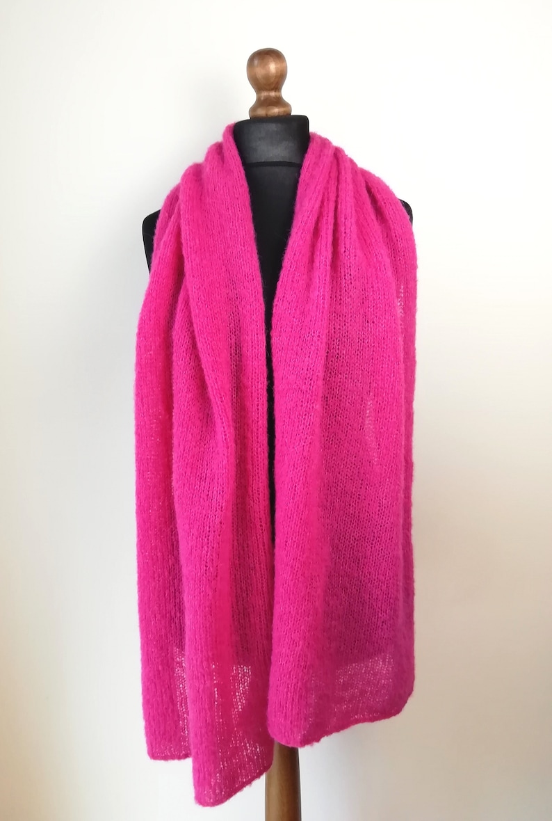 May include: A vibrant, hot pink scarf displayed on a black mannequin head and wooden stand. The scarf is made of a soft, knitted material and drapes down in long, flowing folds. The background is a plain white wall.