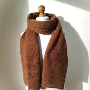 May include: A brown knitted scarf displayed on a white mannequin. The scarf is a solid color, with a thick, textured knit. The scarf is draped around the mannequin's neck and hangs down the front. The scarf appears to be made of a soft, warm material.