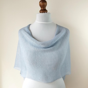 Può includere: Uno scialle in maglia azzurro semitrasparente drappeggiato su un manichino. Il capo presenta una trama fine a maglia aperta con un leggero luccichio, un'ampia scollatura e una silhouette morbida e fluida che copre le spalle e il busto.