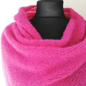 May include: A vibrant fuchsia-colored knitted shawl draped over a black mannequin. The shawl is made of a soft, open-weave material, creating a delicate and airy appearance. The bright pink color is eye-catching and adds a pop of color.