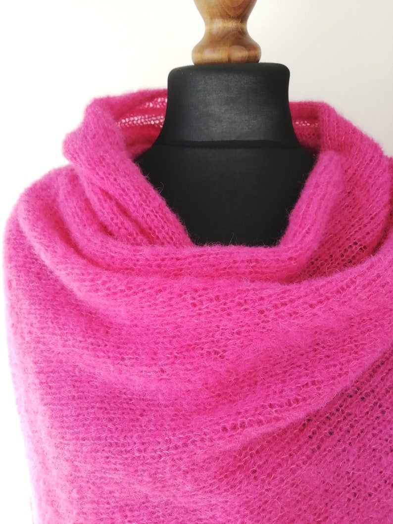 May include: A vibrant fuchsia-colored knitted shawl draped over a black mannequin. The shawl is made of a soft, open-weave material, creating a delicate and airy appearance. The bright pink color is eye-catching and adds a pop of color.
