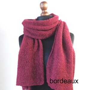 May include: A burgundy-colored knitted scarf draped over a mannequin. The scarf is thick and textured, with a rectangular shape. The word "bordeaux" is printed on the scarf. The scarf is displayed against a white background.