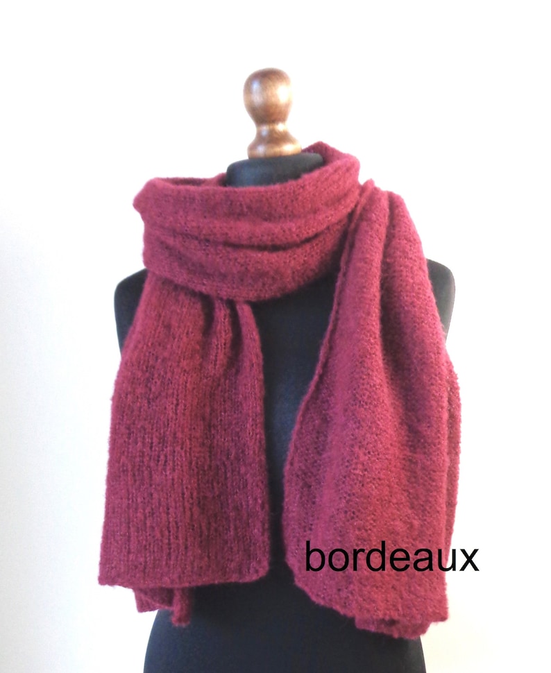 May include: A burgundy-colored knitted scarf draped over a mannequin. The scarf is thick and textured, with a rectangular shape. The word "bordeaux" is printed on the scarf. The scarf is displayed against a white background.