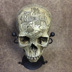 May include: A detailed skull sculpture with the word "Ravenclaw" etched on the forehead. The skull is off-white with dark shading, mounted on a black metal stand. The Ravenclaw crest, featuring a raven, is visible.