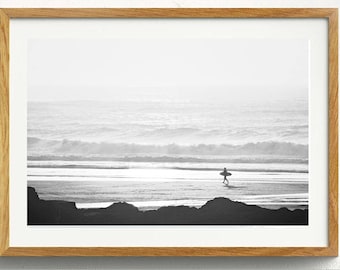 May include: A black and white photograph of a surfer walking on a beach with a surfboard under their arm. The ocean is in the background with waves crashing on the shore.