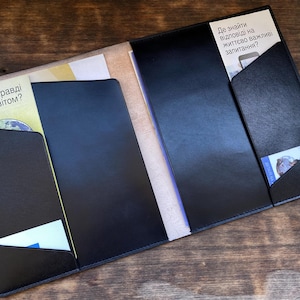 Könnte beinhalten: Ein schwarzer Lederordner mit mehreren Taschen und Klappen. Der Ordner hat ein beigefarbenes Innenleben und ist geöffnet, um die Taschen zu zeigen. Der Text auf dem Ordner lautet "Де знайти відповіді на життєво важливі запитання?" und "Хто насправді керує світом?"