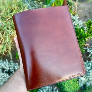 Könnte beinhalten: Ein braunes Leder-Portfolio mit dem Text "Jason Black" auf der unteren rechten Ecke. Das Portfolio wird in einer Hand gehalten, mit einem verschwommenen Hintergrund aus grünem Laub.