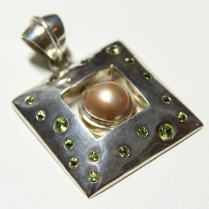 May include: A silver pendant with a square frame and a pearl in the center. The frame is decorated with small green gemstones.