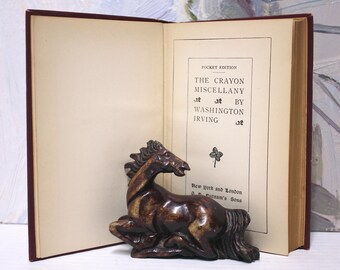May include: An open book titled "The Crayon Miscellany" by Washington Irving, Pocket Edition. The book is resting on a white surface with a small, brown, carved horse figurine in front of it.