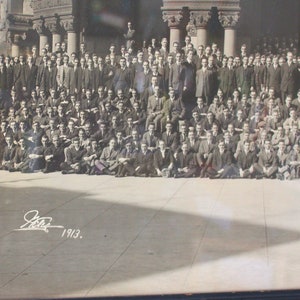 May include: A black and white photograph of a large group of men standing and sitting in front of a building. The men are all wearing suits and ties. The photo is dated 1913.