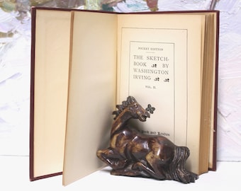 May include: An open book titled "The Sketch-Book by Washington Irving, Vol. II, Pocket Edition" with a brown wooden horse figurine resting on the pages.