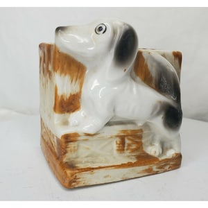 May include: White ceramic dachshund figurine with black markings, sitting on a brown and white ceramic base. The figurine is a planter.